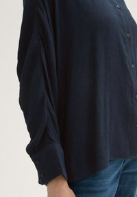 Marineblauwe shirt met een gestructureerde stof, knoopsluiting en opgerolde mouwen. Ontworpen met plooien langs de mouwen en een losse pasvorm.