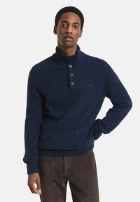 Man in een marineblauwe gestructureerde gebreide trui met een drieknopf ribbelkraag en donkerbruine broek, staand met één hand op de heup.