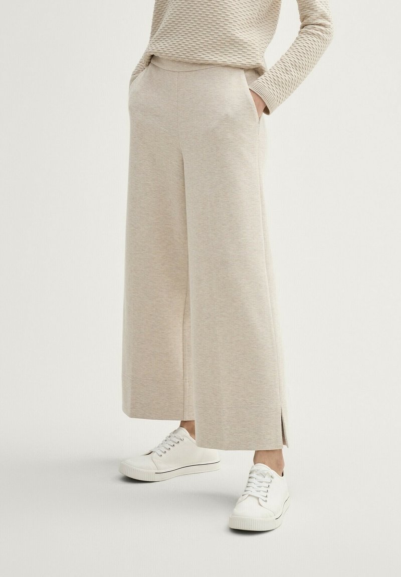 Beiga vida byxor i mjukt tyg, med sidofickor och sprund i fållen, kombinerade med vita sneakers för en avslappnad stil.