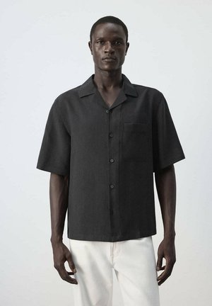 Hombre de pie con una camisa negra de manga corta abotonada y bolsillo en el pecho, y pantalones blancos, sobre un fondo claro y sencillo.