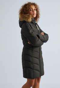 Sieviete ar cirtainiem matiem, tērpta melnā, quiltētā garajā ziemas mētelī ar kažokādas apdari uz kapuces, ar rokām sarautām, noliektā stāvoklī nedaudz šķībi pret gaismu.