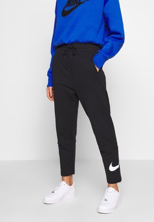 Osoba nosząca czarne spodnie dresowe Nike z białym logo na dolnej części nogawki, białe sneakersy oraz niebieską bluzę Nike z czarnym logo.