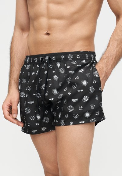 Svarte badeshorts med hvite mønstre av sol, fredstegn, solbriller og kryssede årer, båret av en skjorteløs mann med den ene hånden i lomma.