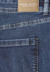 Siniset farkut, joissa on takatasku ja ruskea nahkapatch, jossa lukee "Street One - Denim Atelier, Suunniteltu intohimolla."