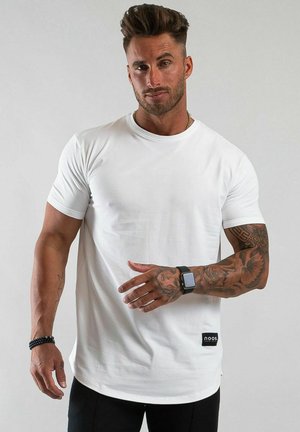 Man med tatueringar bär vit t-shirt, svart smartklocka och armband, står mot en enfärgad ljus bakgrund med neutralt ansiktsuttryck.