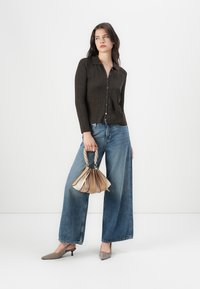 Brun ribbad knäpptröja, blå vida jeans, grå spetsiga klackar, håller en flerfärgad plisserad handväska med flätade handtag.