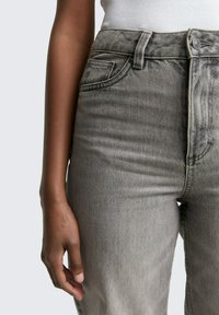 Närbild av en person som bär högtåiga grå denimjeans och en vit ribbad topp, med vänster hand avslappnat vid sin sida.
