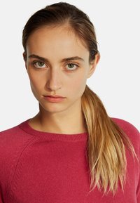 Czerwona sweter z okrągłym dekoltem i teksturowanym materiałem. Modelka ma proste brązowe włosy w kucyku i neutralny makijaż.