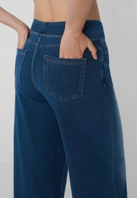 Jeans de mezclilla azul de pierna ancha con una cintura alta, que cuentan con dos bolsillos de parche en la parte posterior, costuras sutiles y una textura de tela suave.