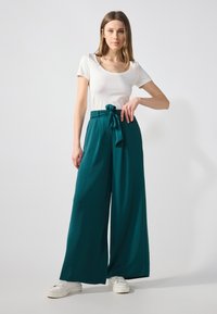 Pantalons larges teal avec une ceinture nouée, en tissu texturé, associés à un haut à manches courtes blanc, le tout porté avec des baskets blanches.
