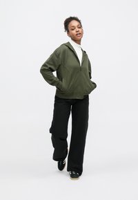 Olívazöld, cipzáras kapucnis pulóver testhez simuló szabással, elülső zsebekkel és puha tapintású anyagból, fekete bő szárú nadrággal és fekete tornacipővel párosítva.