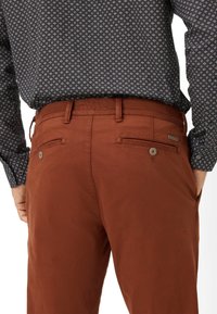 Braune Baumwollhosen mit glatter Textur, ausgestattet mit zwei Gesäßtaschen mit Knopfverschlüssen und einem Gürtelschlaufen-Design.