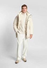 Giacca a vento di colore crema con colletto alto e chiusura a zip, abbinata a un maglione crema a treccia e pantaloni bianchi sartoriali. Sneakers marroni.