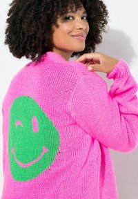 Maglione lavorato a maglia rosa con un design di faccina sorridente verde sulla schiena. Presenta maniche lunghe e una vestibilità morbida.