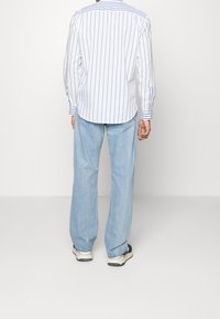 Person klädd i en vit knäppt skjorta med vertikala blå ränder, ljusblå jeans och vita sneakers, står vänd bort på en enfärgad bakgrund.