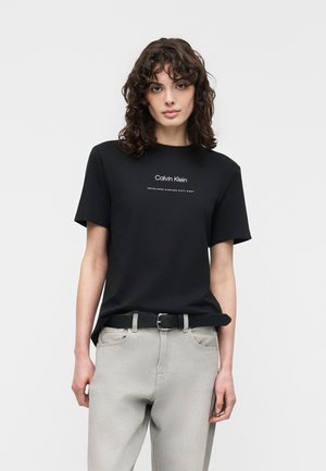 Γυναίκα με σγουρά σκουρόχρωμα μαλλιά, φορώντας μαύρο T-shirt Calvin Klein και ανοιχτό γκρι τζιν με μαύρη ζώνη, στέκεται μπροστά σε απλό φόντο.