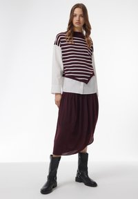 Pull rayé marron et blanc sur une chemise blanche, associé à une jupe longue marron et des bottes noires jusqu'au genou avec des boucles et un talon épais.
