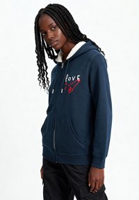 Gilet à capuche zippé marine avec un graphique "love" en blanc et rouge, comprenant une capuche douce et doublée en polaire ainsi que des poches frontales, au design décontracté.