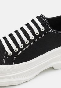 Schwarze Leinensneaker mit weißen Schnürsenkeln, Gummisohle und kontrastierender weißer Naht. Hat runde Ösen und eine strukturierte Seite.