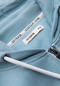 Col de sweat à capuche bleu clair avec étiquette en tissu « ombre cotton line », cordon blanc et tirette de fermeture éclair noire visibles.