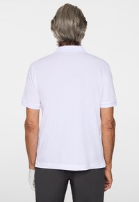 Herrar vit pikétröja med krage, korta ärmar, strukturerad tyg och rak nederkant. Anmärkningsvärt detaljer: en hand bär en handske.