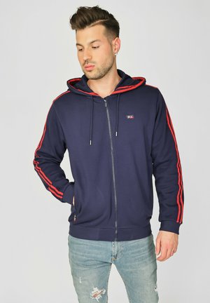 Marineblå zip-up hættetrøje i bomuld med røde sidestropper, snoretræk i hætten, frontlommer og logo-patch på brystet. Båret med slidte blå jeans.