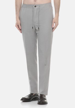 Uomo che indossa pantaloni grigio chiaro su misura con bottone e coulisse in vita, abbinati a mocassini in pelle nera, in piedi su sfondo bianco.