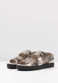 Kermavahti ja beez sandaal, millel on nahkakirju muster, kaks laia rihma, avatud varvastega disain ja must kummitald. Reguleeritav pahkluu klambriga detail.