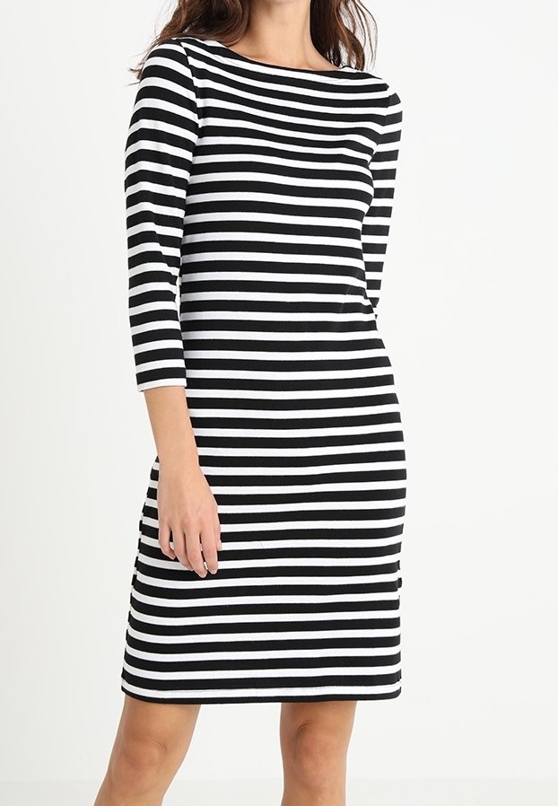Femme portant une robe noire et blanche à rayures horizontales jusqu'aux genoux avec des manches trois-quarts sur un fond uni.