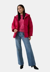 Giacca a vento rosa con motivo trapuntato, abbinata a un maglione rosa coordinato e jeans a gamba larga blu. Stivaletti neri completano l'outfit.