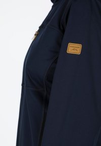 Marinejacke aus weichem Material, mit einer Reißverschlusstasche und einem beigen Markenpatch auf dem Ärmel. Texturierte Nähte verleihen Detail.