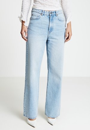 Jean large à taille haute bleu clair avec une finition délavée, doté de poches avant et d'accents en métal, associé à des chaussures blanches pointues.