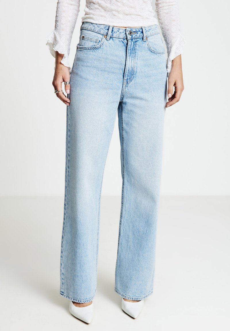 Jeans azzurri chiaro a vita alta con gamba larga e finitura sbiadita, caratterizzati da tasche frontali e dettagli in metallo, abbinati a scarpe bianche a punta.