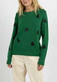 Pull vert en matériau doux avec un design classique. Présente des motifs de cœurs noirs scintillants. Manches longues, coupe décontractée.