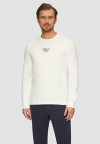 Witte lange mouw t-shirt van katoen, met een ronde hals en een bedrukte logo op de borst. Gepaard met donkere broeken.