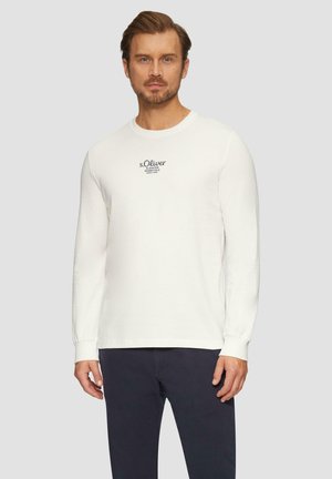 Weißes Langarm-T-Shirt aus Baumwolle mit Rundhalsausschnitt und einem bedruckten Logo auf der Brust. Kombiniert mit dunklen Hosen.