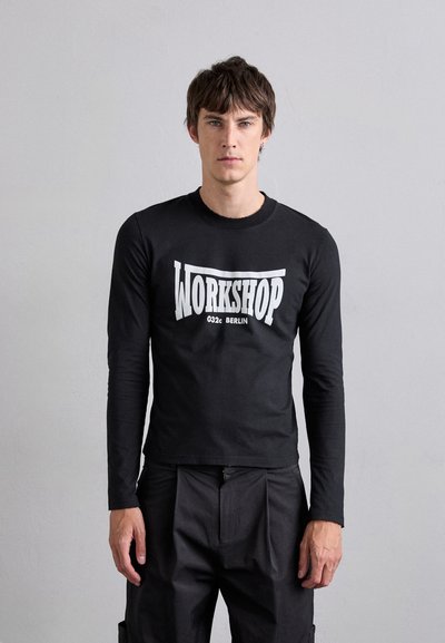 Schwarz longsleeve T-Shirt aus glattem Baumwollmaterial mit einem auffälligen weißen "WORKSHOP"-Grafik und dem Text "032c BERLIN" auf der Vorderseite. Enganliegendes Design.