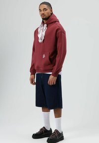 Donkerrood capuchontrui met een voorzak, gedragen over een wit shirt. Gecombineerd met denim shorts tot op de knie en zwarte schoenen met roze veters.