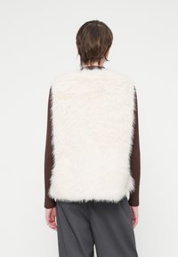 Kamizelka z sztucznego futra w odcieniu off-white, charakteryzująca się miękką teksturą, wysokim kołnierzem i bezrękawowym krojem, noszona na brązowej bluzce z długim rękawem.