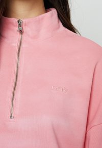 Ljusrosa pullover med kvartszipp i mjukt tyg, med ribbad struktur och en liten broderad Levi's-logotyp på bröstet.