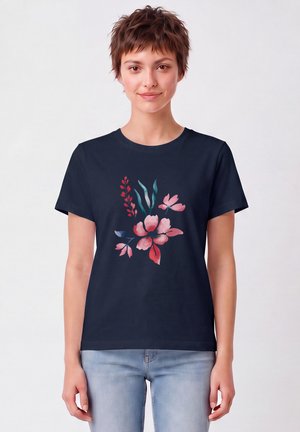 Jeune femme aux cheveux courts et bruns portant un t-shirt bleu marine avec un motif floral rouge et rose, associé à un jean bleu clair, debout sur un fond blanc.