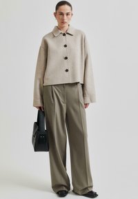 Bežová zkrácená bunda s černými knoflíky, spojená s olivově zelenými širokými kalhotami. Modelka nese černou tašku obdélníkového tvaru.