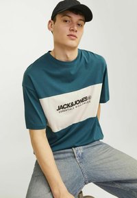 Turquoise og creme t-shirt med korte ærmer, der har et vandret logo design på brystet. Kombineret med lyseblå jeans.