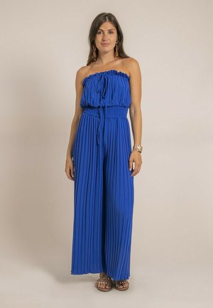 Femme portant une combinaison bleu roi plissée sans bretelles, debout les mains détendues, portant des boucles d'oreilles, un bracelet et des sandales cloutées.