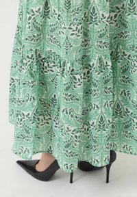 Robe à motifs verts et blancs avec des dessins de feuilles, des couches superposées et une silhouette fluide, portée avec des talons aiguilles noirs.