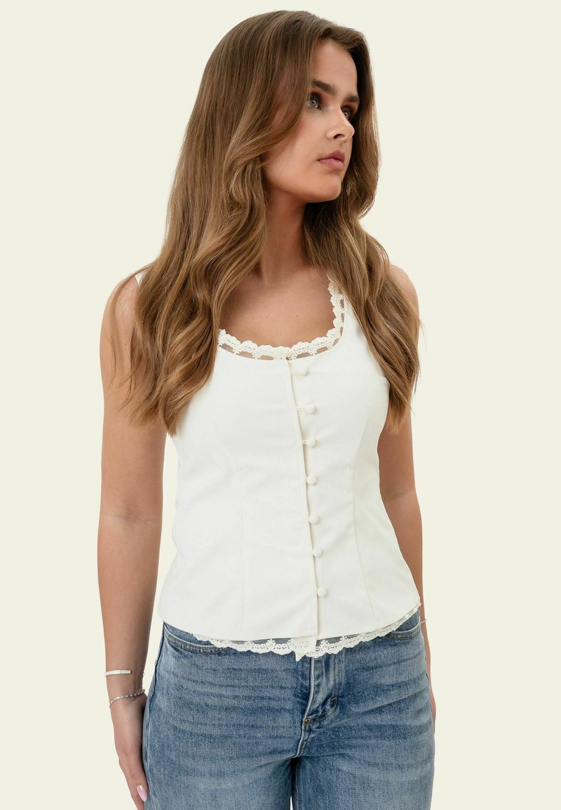 Femme aux longs cheveux ondulés portant un top blanc sans manches boutonné avec bordure en dentelle et un jean bleu, regardant sur le côté.