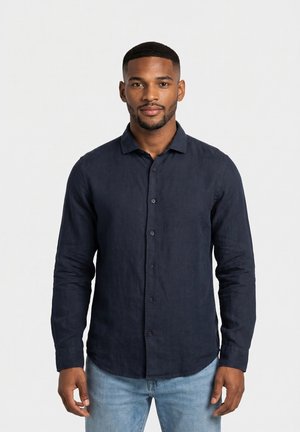 Homme aux cheveux courts et à la barbe, portant une chemise sombre boutonnée et un jean bleu clair, debout devant un fond blanc uni.