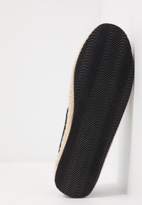 Semelle de chaussure noire avec un motif texturé ondulé, bord tressé beige clair, vue de dessous sur fond blanc.