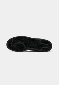 Semelle d'une chaussure noire montrant un motif de semelle zigzag avec des rainures courbes et droites sur un fond blanc.