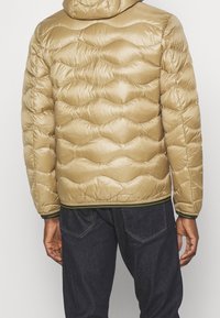 Giacca imbottita beige con cappuccio, caratterizzata da un motivo a onde trapuntate, polsini elasticizzati, orlo verde a contrasto e una texture liscia e lucida.